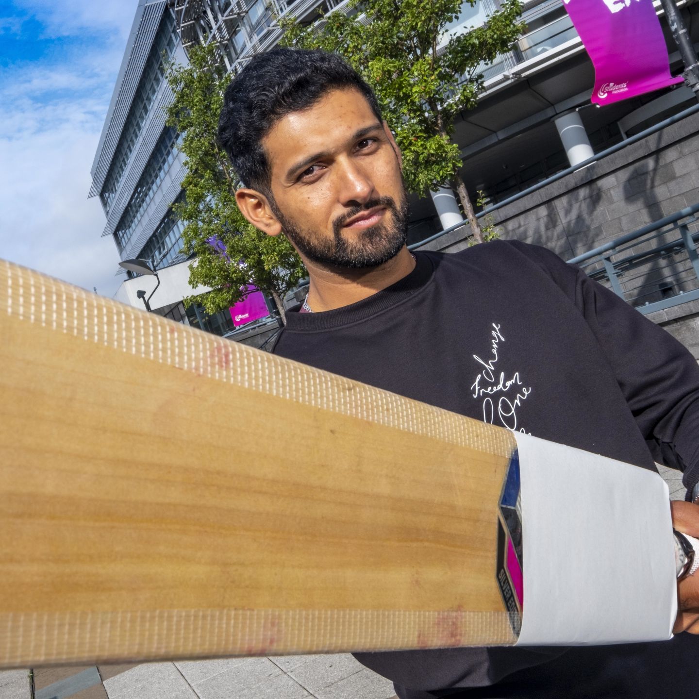 Sikandar Raza, a GCU alumnus and an international cricketer, holding a cricket bat while standing in the exterior of Glasgow campus.