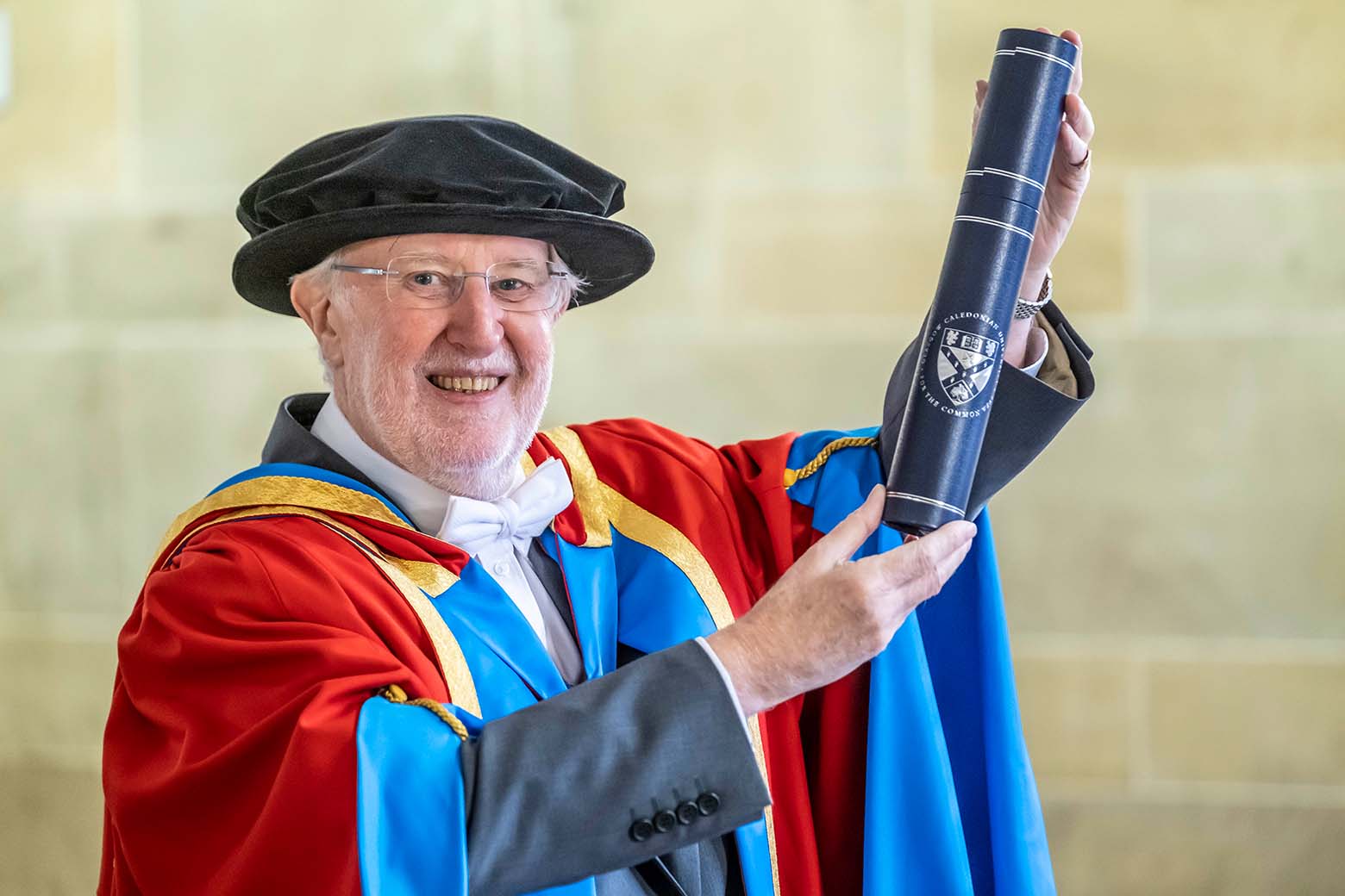 GCU Honorary Graduate Dr Ian Young wearing his graduation hood and hat and holding a degree container.