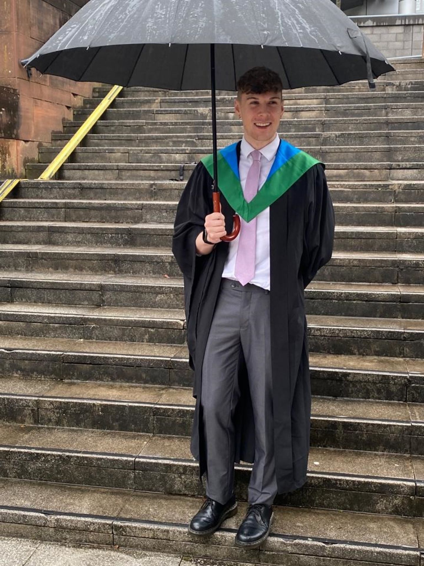 GCU Alumnus Shaun Greenhorn in Graduation gown and robe, holding an umbrella.