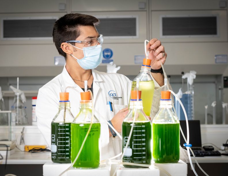 A young male student with short dark hair wearing a facemask and goggles inspects a beaker of pale yellow liquid in a lab. They are surrounded by measuring beakers filled with bright green liquid.