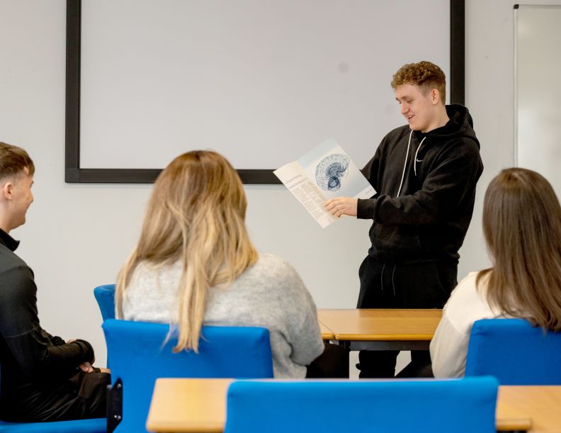 A young male student stands at the front of a classroom with a blue psychology booklet with a brain illustration on the front..