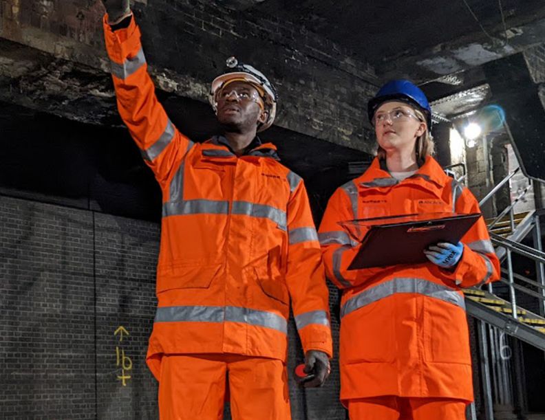 Two students in bright orange hi-vis jumpsuits and helmets are inspecting underneath a bridge. On the left, a young black male points upwards and on the right a young white female looks up while holding a clipboard.