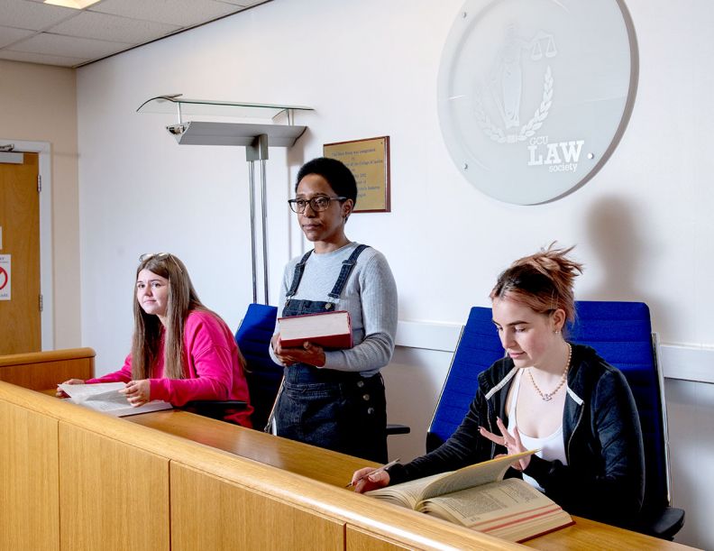 A young black female student with short black hair and large black glasses stands up in a moot room stand holding a book. To their left, a young white female with long brown hair looks out into the moot room and holds open a book. To their right, a young white female with pink-brown hair flips through a book.