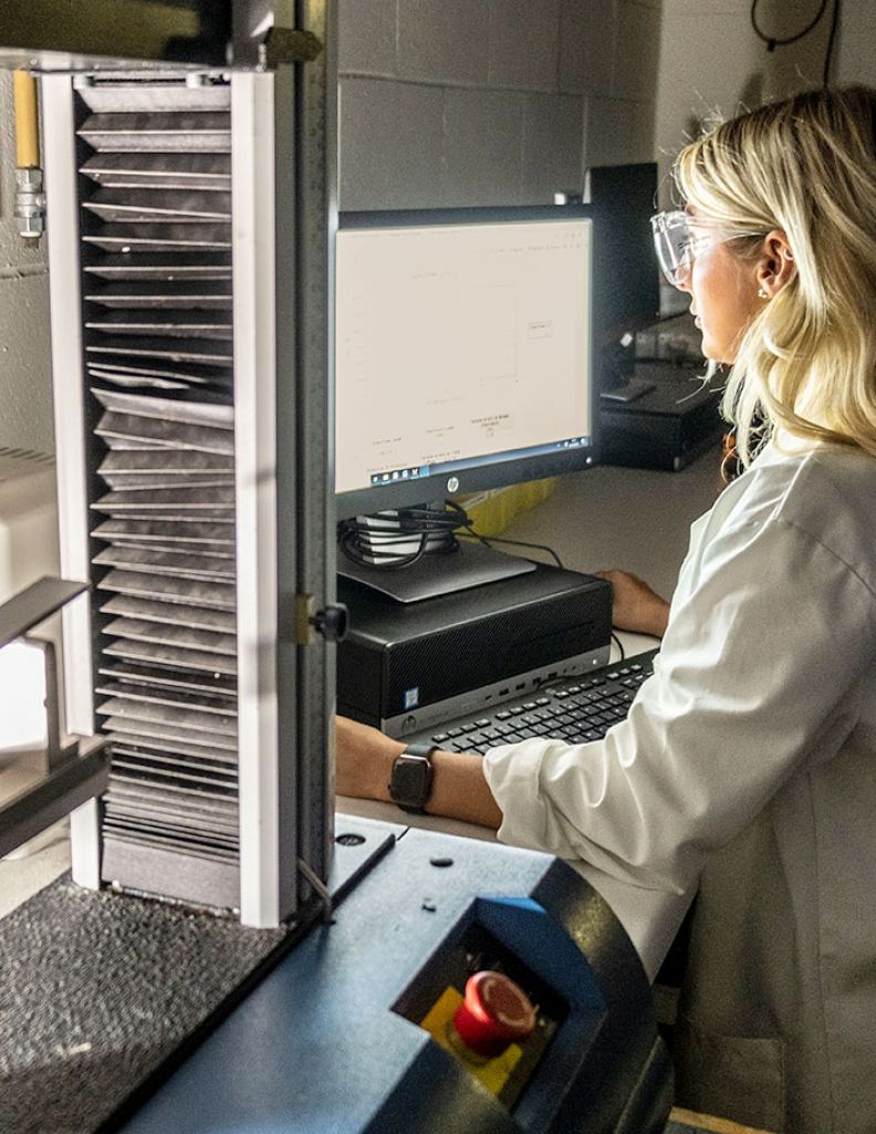 A young white female student in lab goggles and lab coat uses a computer-assisted engineering machine.