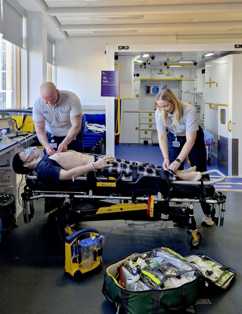 Two nursing students, one male, one female, perform CPR on a mannequin on a gurney. A simulation ambulance is in the background.