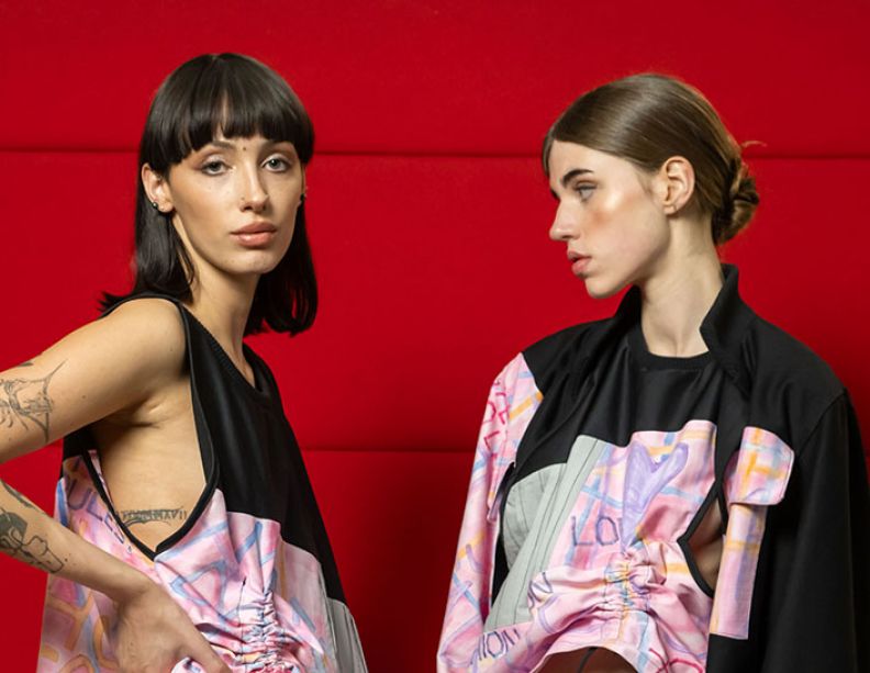 Two female models wearing a student's black and pink upcycled fashion creation. The model on the left is mixed race with a brown bob and fringe looking at the camera and the model on the right is white with light-brown hair in a bun and looking off to the left.