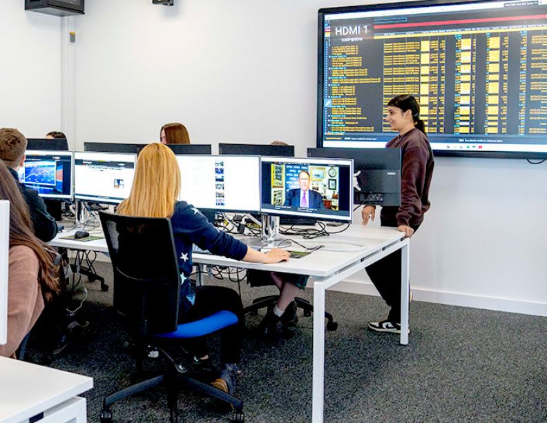 A group of students sit at computers in the Bloomberg finance suite. Behind them are screens which are black screens flashing up economics figures in orange text.