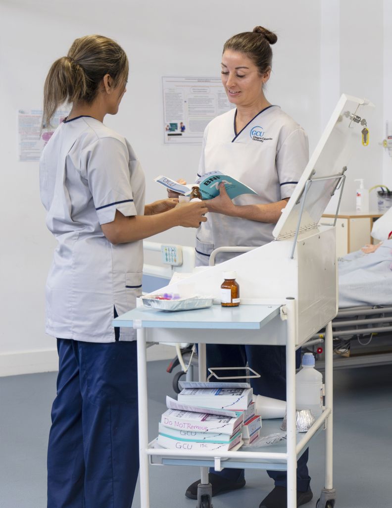 Two female nursing students are standing by a mobile cabinet discussing medication bottles.