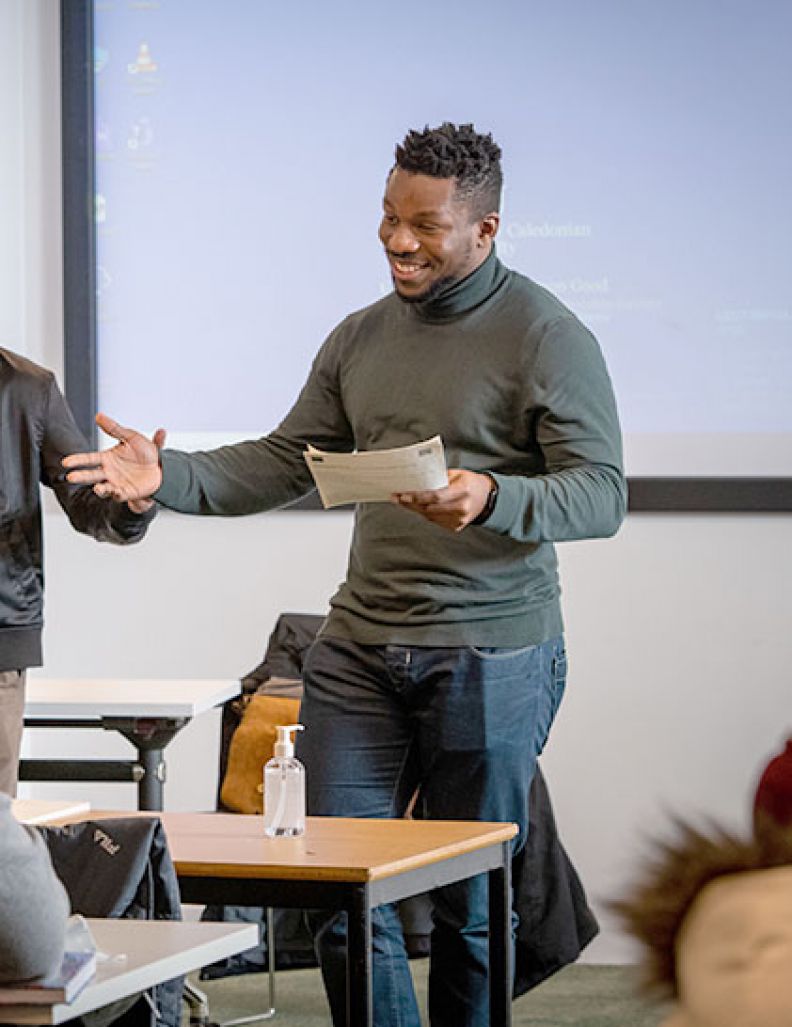 A young black male student with short black hair stands at the front of a room holding a piece of paper. They are giving a presentation with a big smile.